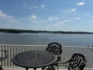 Un balcon avec une table ronde et des chaises, donnant sur une étendue d’eau, avec une terre lointaine et des arbres visibles au loin.