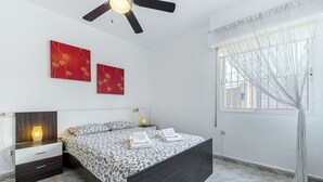 Un dormitorio con cama, mesitas de noche, ventilador de techo, obra de arte en la pared y ventana con cortinas.