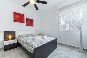 A bedroom with a bed, bedside tables, a ceiling fan, artwork on the wall, and a window with curtains. - Apartment Ground floor Doña Ines Torrevieja (Torrevieja)