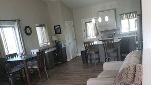 Ett rymligt vardagsrum med en soffa, ett matbord och stolar, ett kök i bakgrunden och ett stort fönster som släpper in naturligt ljus.