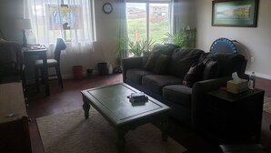 Ett vardagsrum med en soffa, ett soffbord och en matplats med ett bord och stolar. Det finns fönster med gardiner, en klocka på väggen och en inramad tavla.