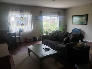 Ein Wohnzimmer mit einer Couch, einem Couchtisch und einem Essbereich mit Tisch und Stühlen. Es gibt Fenster mit Vorhängen, eine Uhr an der Wand und ein gerahmtes Bild.