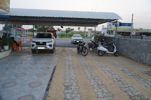 A covered parking area with a paved walkway, a white SUV, and several motorcycles parked. - Hotel Radha Raman and Restaurant (Danta Ramgarh)