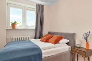 Ein Schlafzimmer mit Bett, Nachttisch, einem Fenster mit Vorhängen, Heizkörper und einer Zimmerpflanze.
