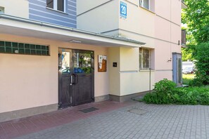 Ein Gebäudeeingang mit einer Metalttür, einem Schild mit der Zimmernummer 20 und einem kleinen, vergitterten Fenster.