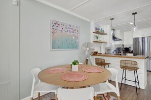 A modern kitchen with white cabinetry, a central island, and a dining area with a round table and white chairs. - Walking On Sunshine: 2BR Home (Tybee Island)