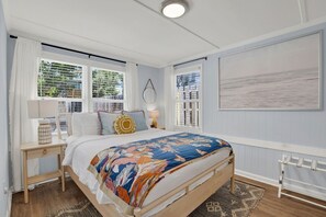 A bedroom with a bed, two bedside tables, a window with blinds, a radiator, and a painting on the wall. - Walking On Sunshine: 2BR Home (Tybee Island)
