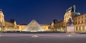 A large, illuminated building with a prominent pyramid-like structure in the center, surrounded by classical architecture. - Palais Royal - Prestige between the Louvre and the Opera (Paris)