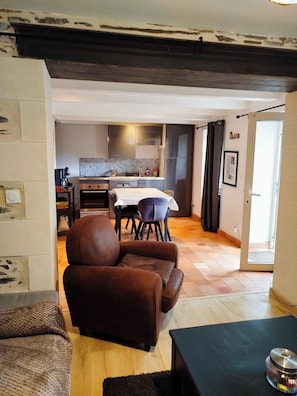 A living room with a brown leather sofa, a dining table with chairs, a kitchen area with cabinets and appliances, and a door leading to another room.