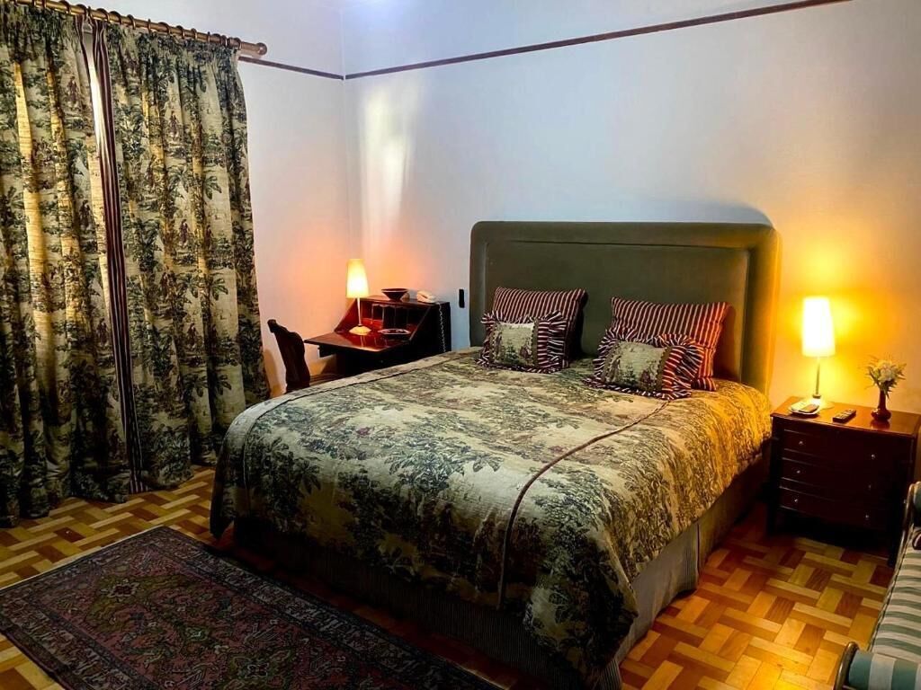 Un dormitorio con cama, dos mesitas de noche, una lámpara, una silla y una alfombra estampada.