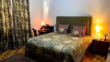Un dormitorio con cama, dos mesitas de noche, una lámpara, una silla y una alfombra estampada.