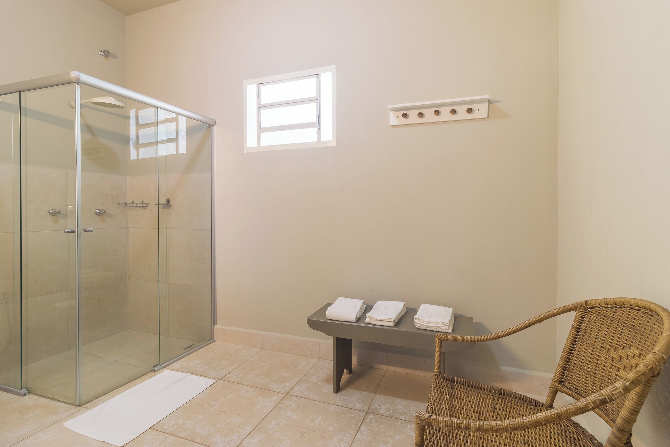 A glass-enclosed shower, a wicker chair, a small table with towels, a window, and a light switch on the wall.