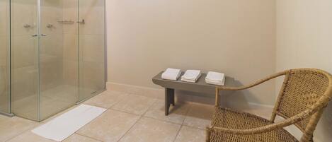 A glass-enclosed shower, a wicker chair, a small table with towels, a window, and a light switch on the wall.