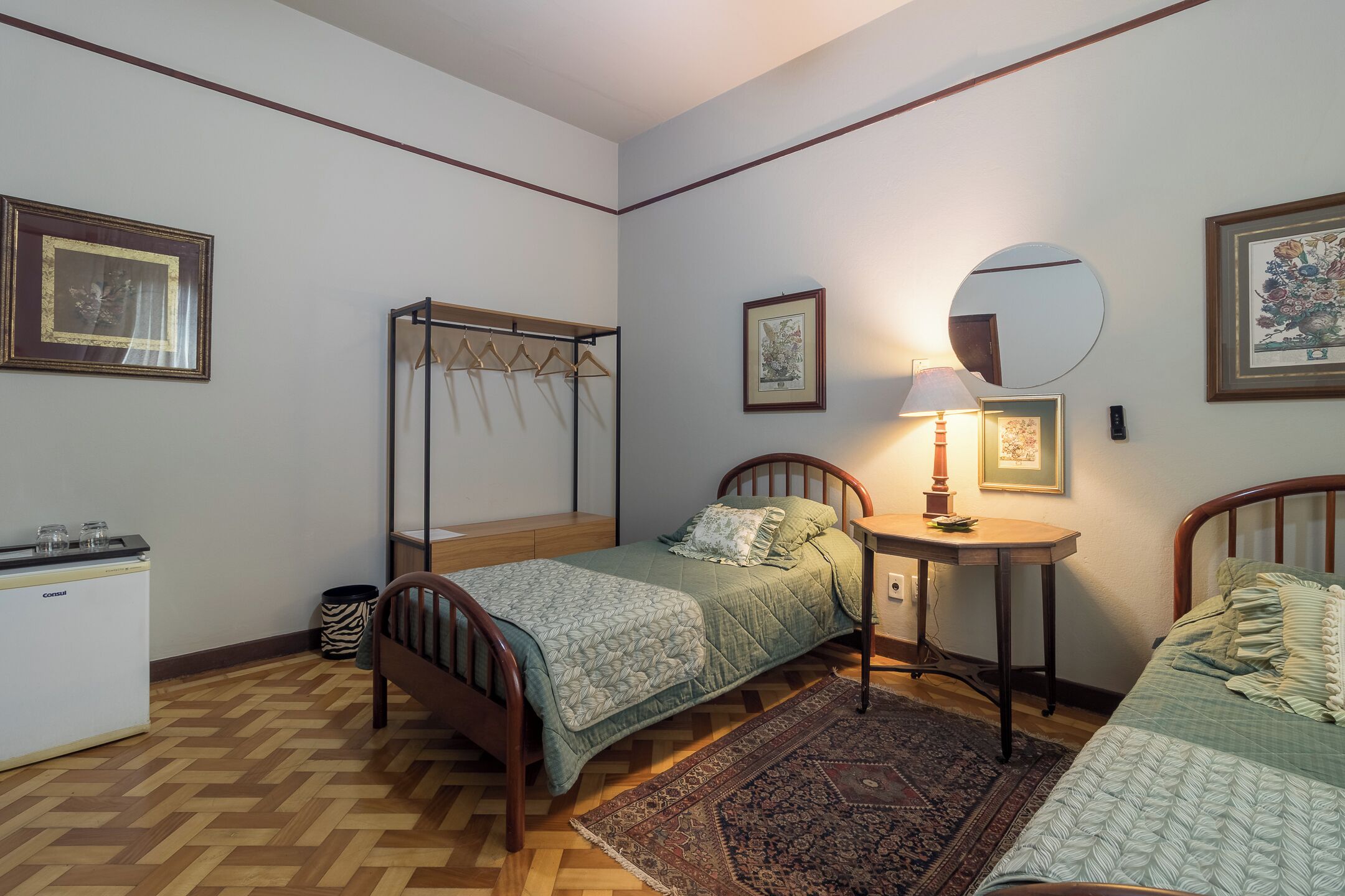 A bedroom with two beds, a bedside table with a lamp, a wardrobe, and a small desk.