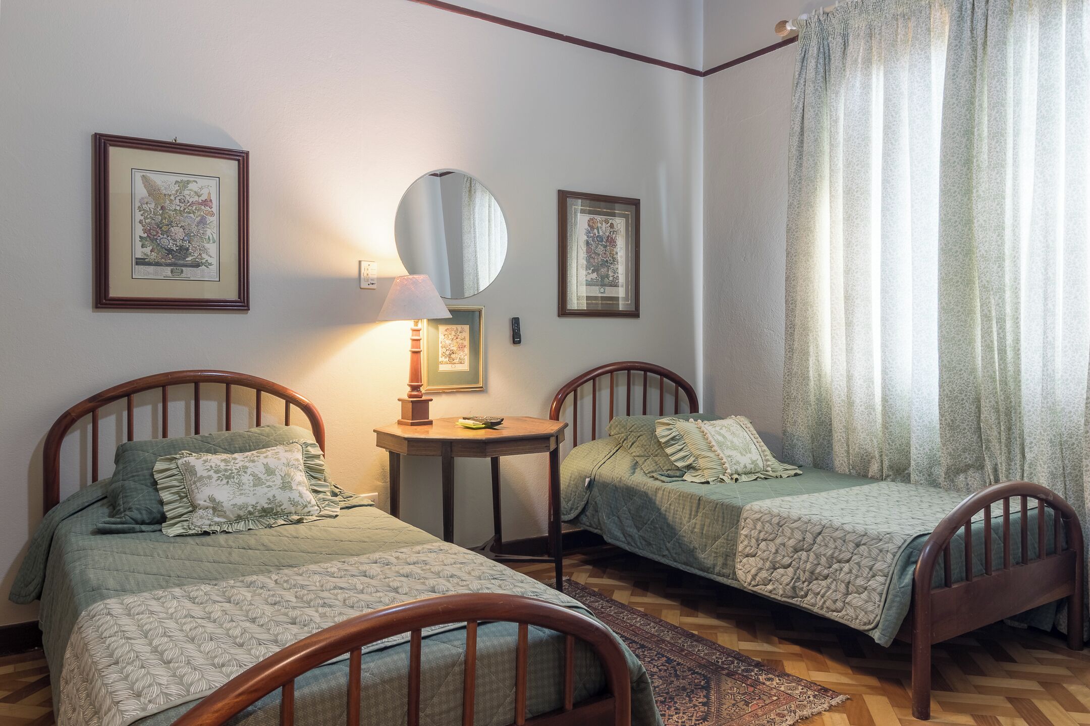 A room with two wooden beds, a round mirror, framed artwork on the wall, and a window with curtains.