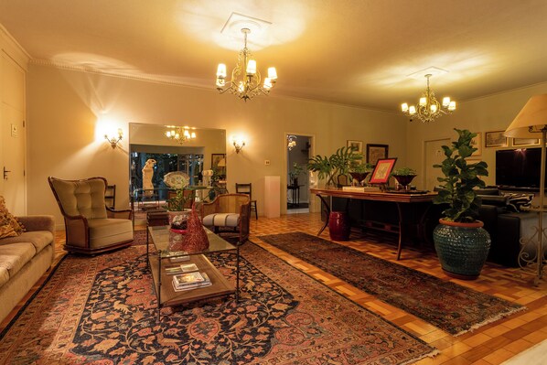 A living room with a sofa, armchairs, a coffee table, a chandelier, a dining table, and a large potted plant. - Pousada Villa do Conde (Brotas)