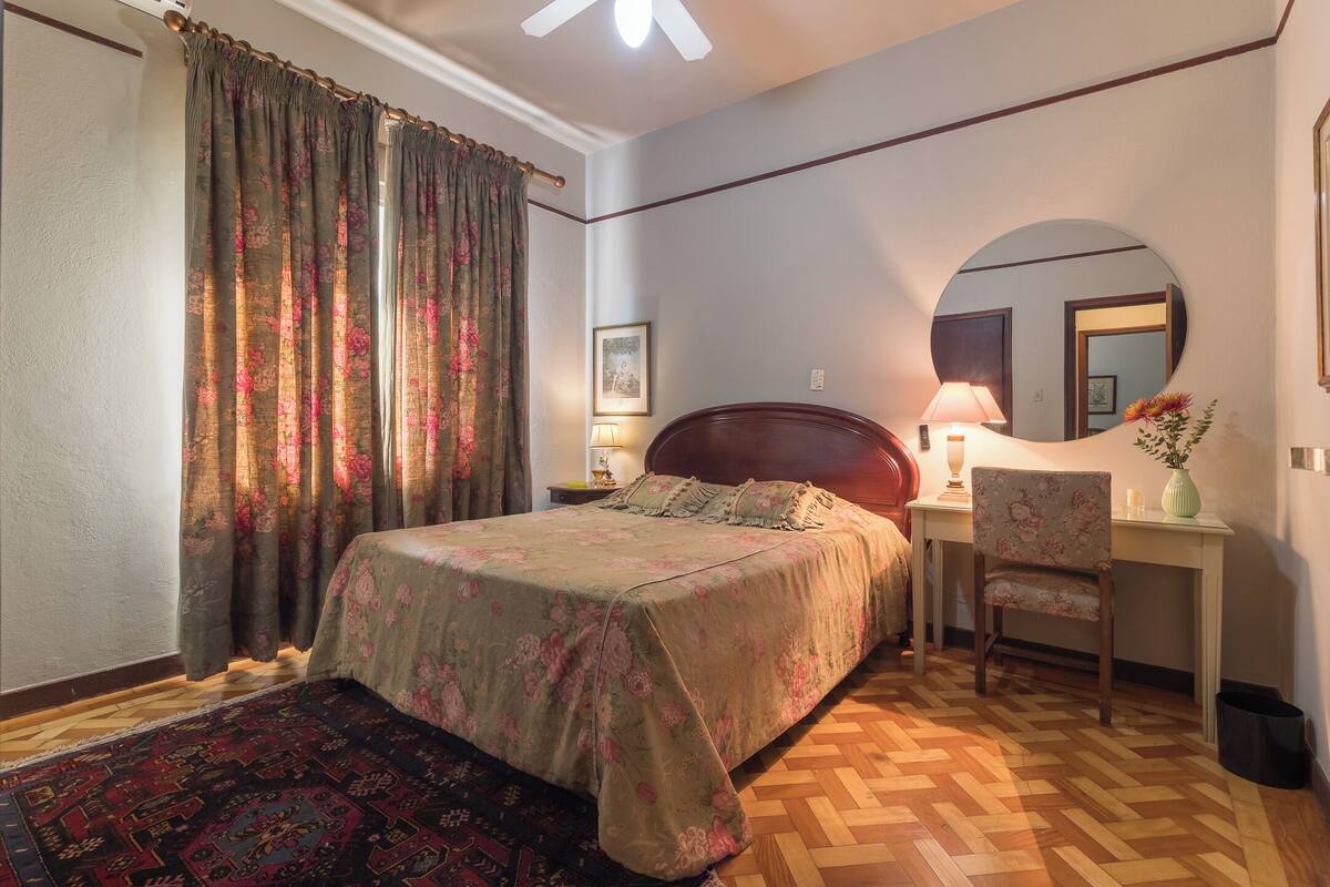 A bedroom with a bed, a desk, a chair, a mirror, and a rug.