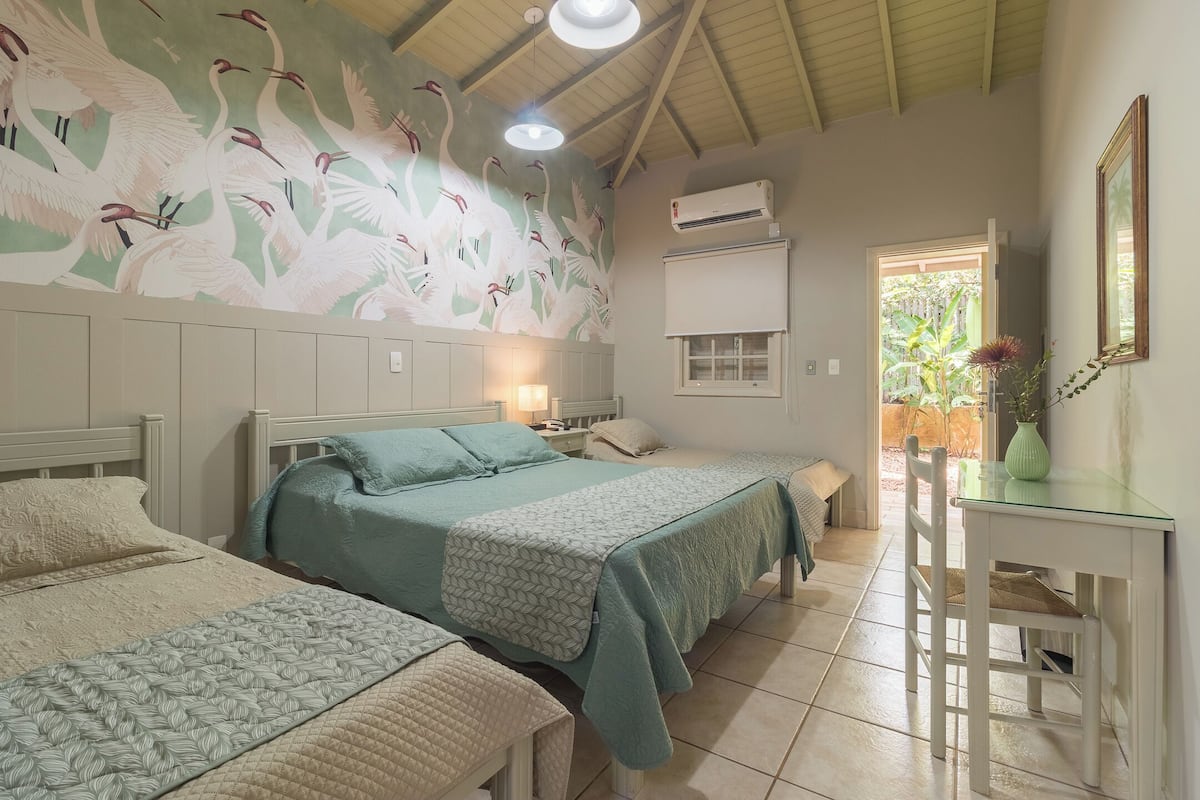 A room with two beds, a desk, a wall-mounted air conditioner, and a window with a view of greenery.
