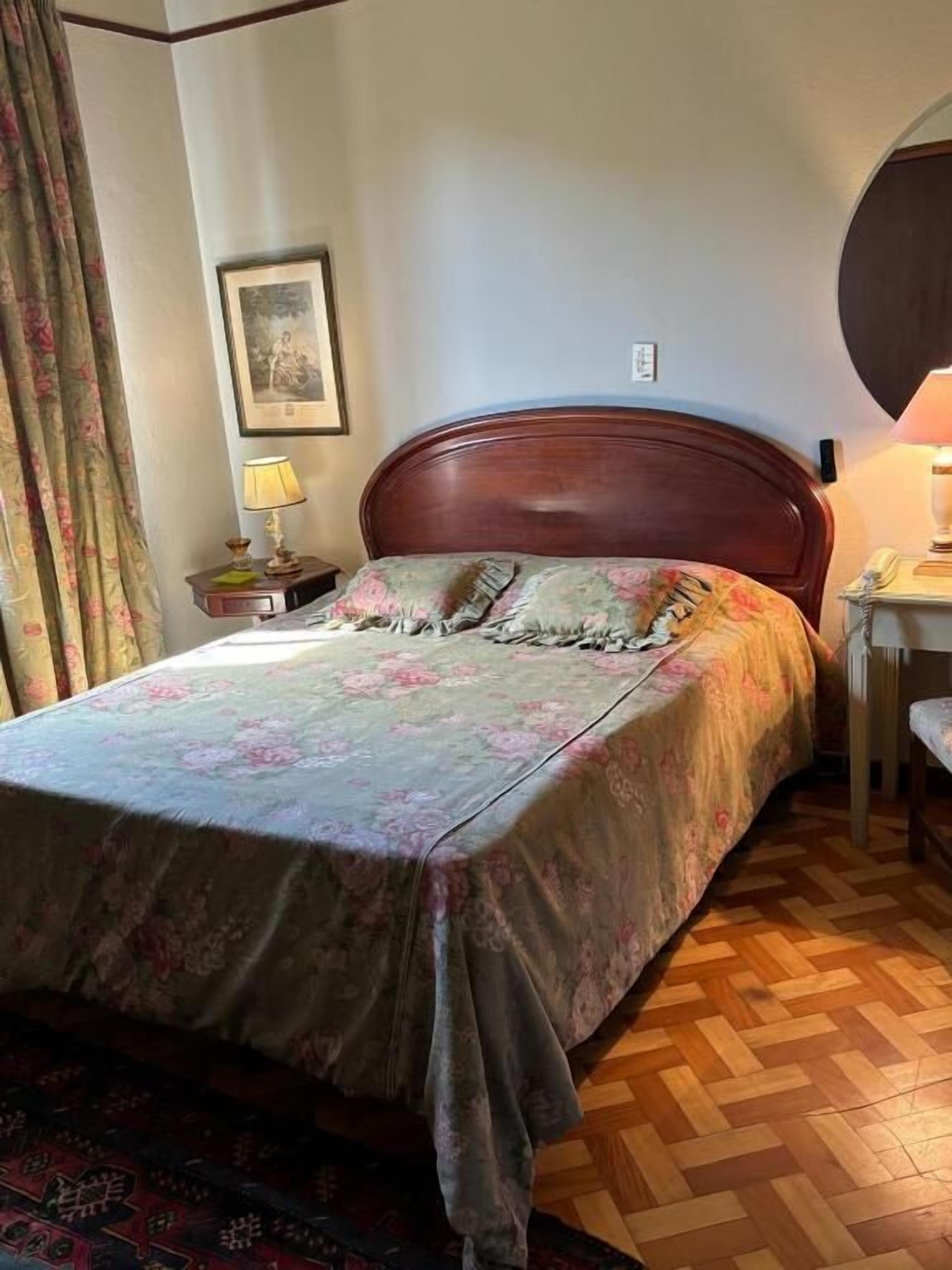 Ein Schlafzimmer mit einem hölzernen Bett, floralem Bettbezug, einem Nachttisch mit Lampe und einem gemusterten Teppich.