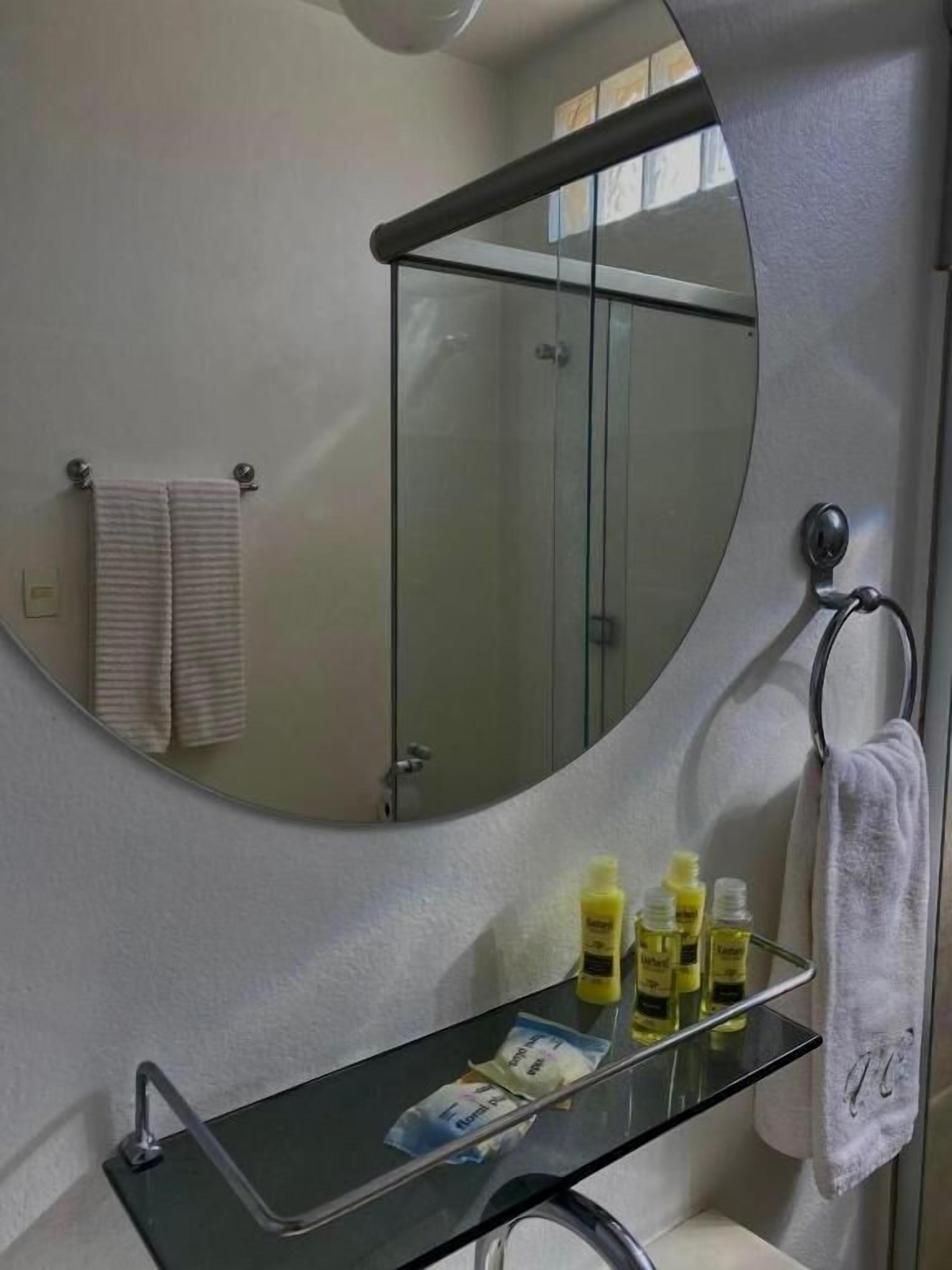 A bathroom with a glass shower enclosure, a round mirror, a glass shelf with toiletries, and a towel rack.