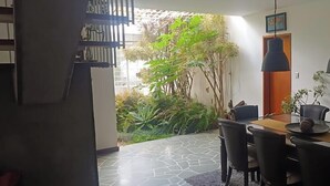 Comedor con una mesa y sillas, una lámpara colgante y vistas a un jardín con diversas plantas.