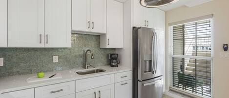 Una cocina con gabinetes blancos, un refrigerador de acero inoxidable, un lavamanos y una ventana con persianas.