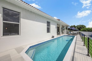 Uma piscina com borda azulejada, um edifício branco com janelas e um céu claro.