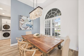A dining area with a wooden table, wicker chairs, a chandelier, a painting of seashells, and a window with blinds. - Coastal Modern Home | Walk To Beach, Food & Shopping (Marco Island)