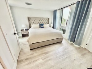 Ein Schlafzimmer mit einem großen Bett, einem Kopfteil, einem Nachttisch, einer Lampe und einem Fenster mit Vorhängen.