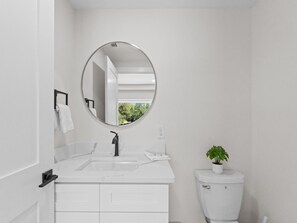 Uma casa de banho moderna com uma pia branca, um espelho redondo, uma sanita e uma planta num vaso.