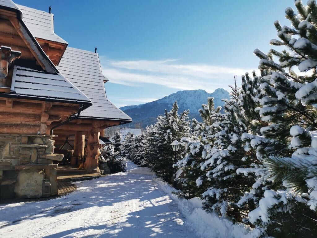 A log cabin with a stone foundation, a wooden porch, and a snow-covered landscape with mountains in the background.