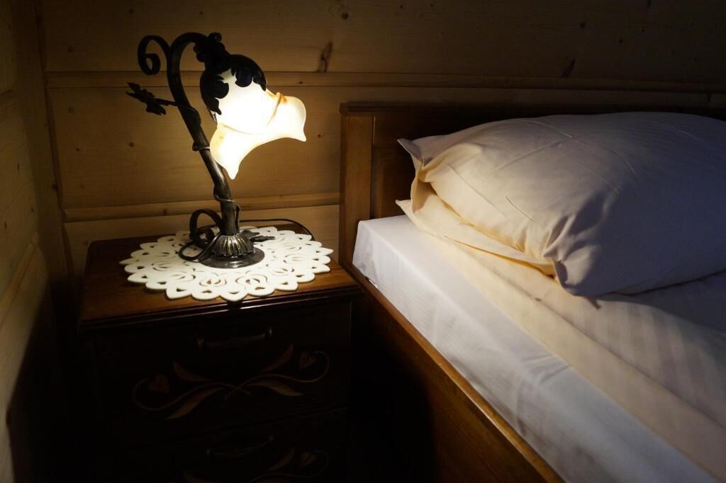 A wooden bedside table with a decorative lamp, a white doily, and a neatly made bed with white pillows.
