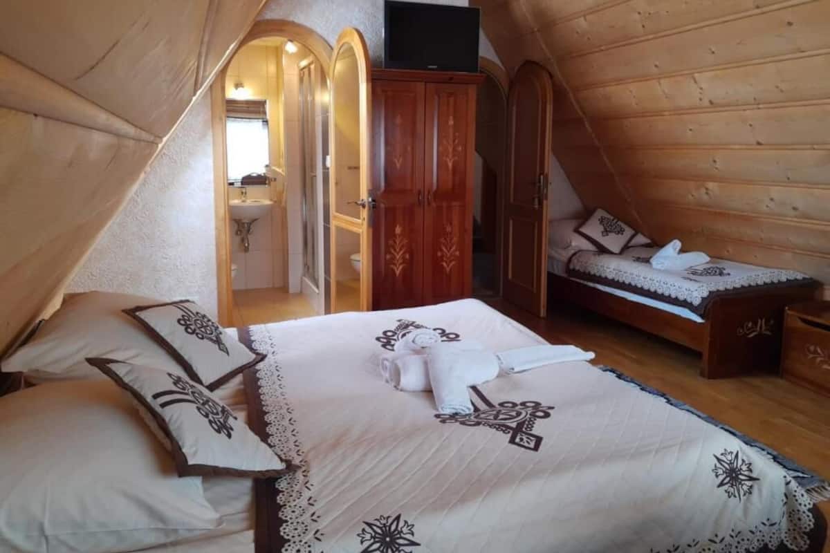 Un dormitorio acogedor en el ático, con techo inclinado de madera, una cama con cubierta de encaje, una mesita de noche pequeña y un armario de madera.