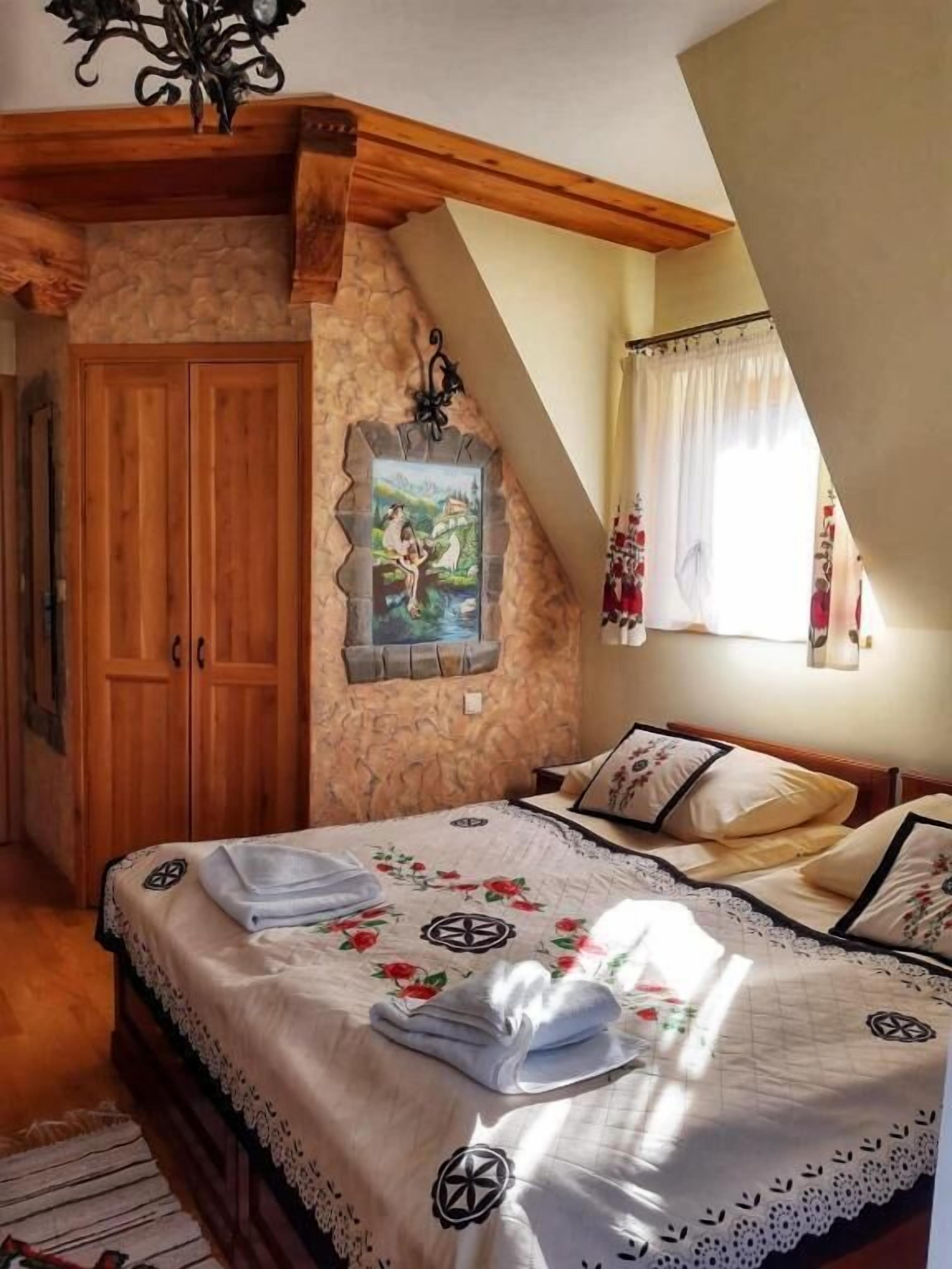 Un dormitorio con pared de piedra, vigas de madera en el techo, una cama con ropa de cama floral, un cuadro y una ventana con cortinas.