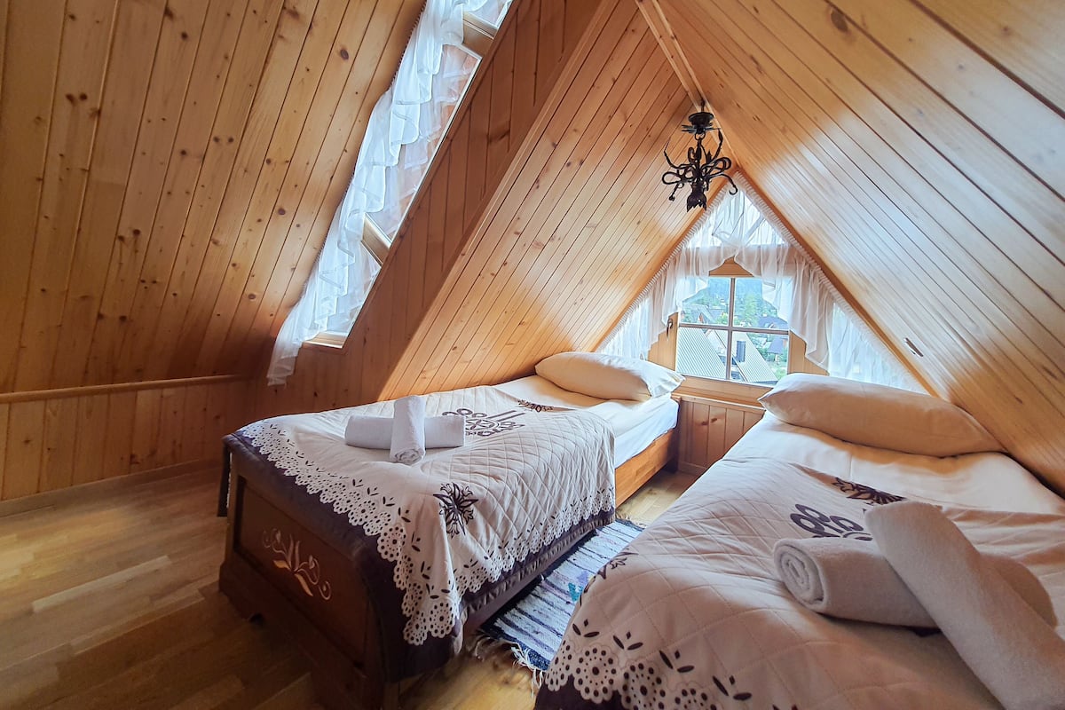Un dormitorio abuhardillado de madera con dos camas, una araña y vistas a casas a través de una ventana.