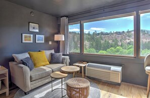 A modern living room with a sofa, a small table, a lamp, and a large window with a view of a mountainous landscape. - New Listing! Cliffside Studio | AC, Pool, Hot Tub, Golf & Ski (Durango)