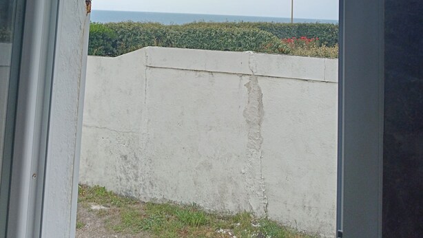 Una puerta abierta que da a un balcón con vistas a un paisaje lejano, un muro de concreto y una farola.