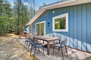 A blue building with a patio area featuring a table and chairs, a grill, and a view of the surrounding forest.