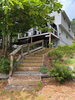 A wooden staircase leading to a house with a balcony, surrounded by trees and a garden.