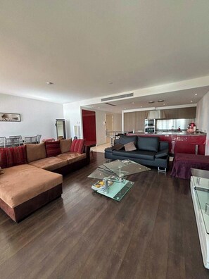 Ein modernes Wohnzimmer mit einer Eckcouch, einem Glastisch und einer Küchenzeile im Hintergrund.