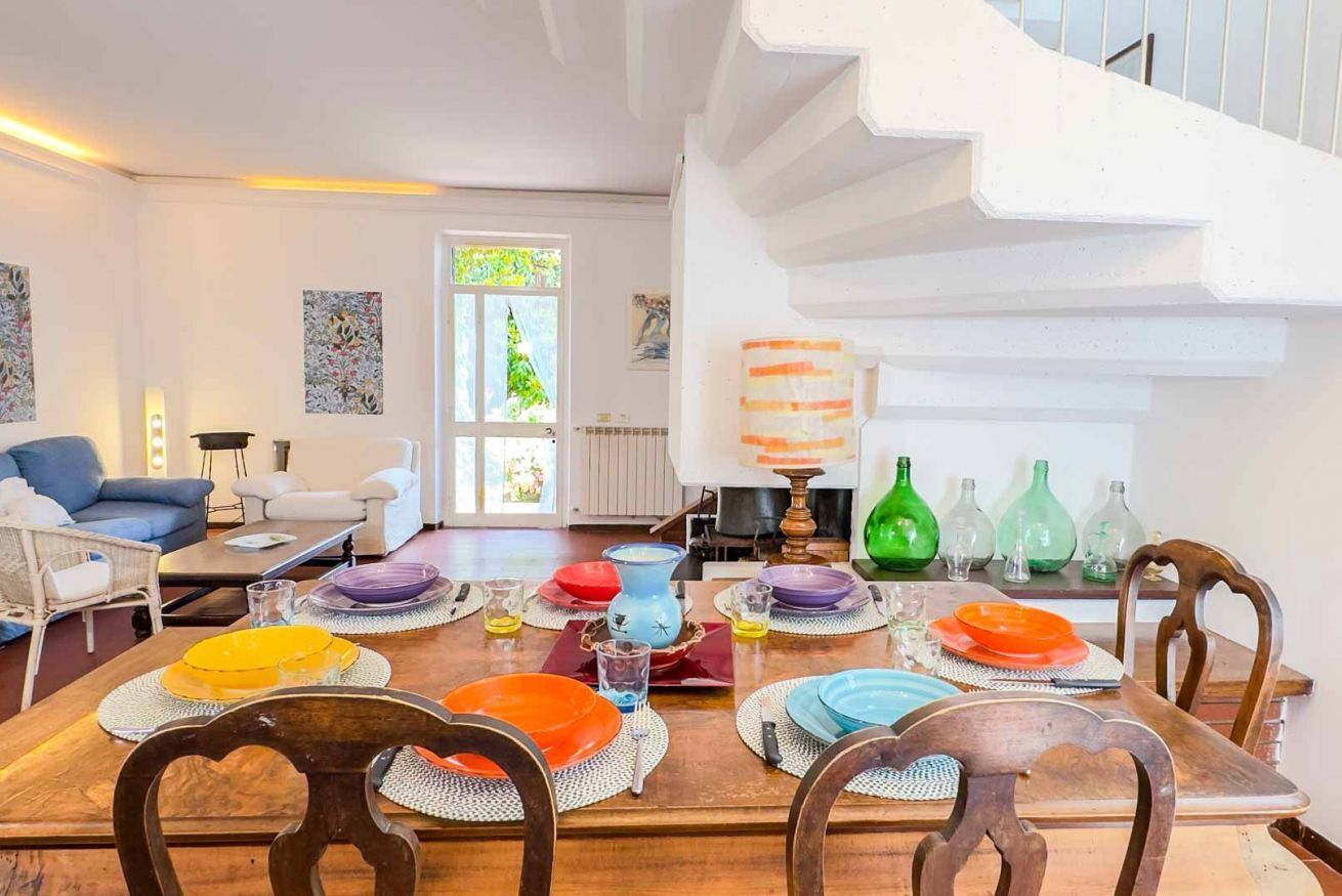 A dining area with a wooden table, chairs, and colorful plates. A staircase is visible in the background.