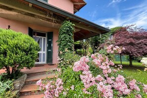 Una casa con porche cubierto, un pequeño jardín de flores rosas y un árbol verde.
