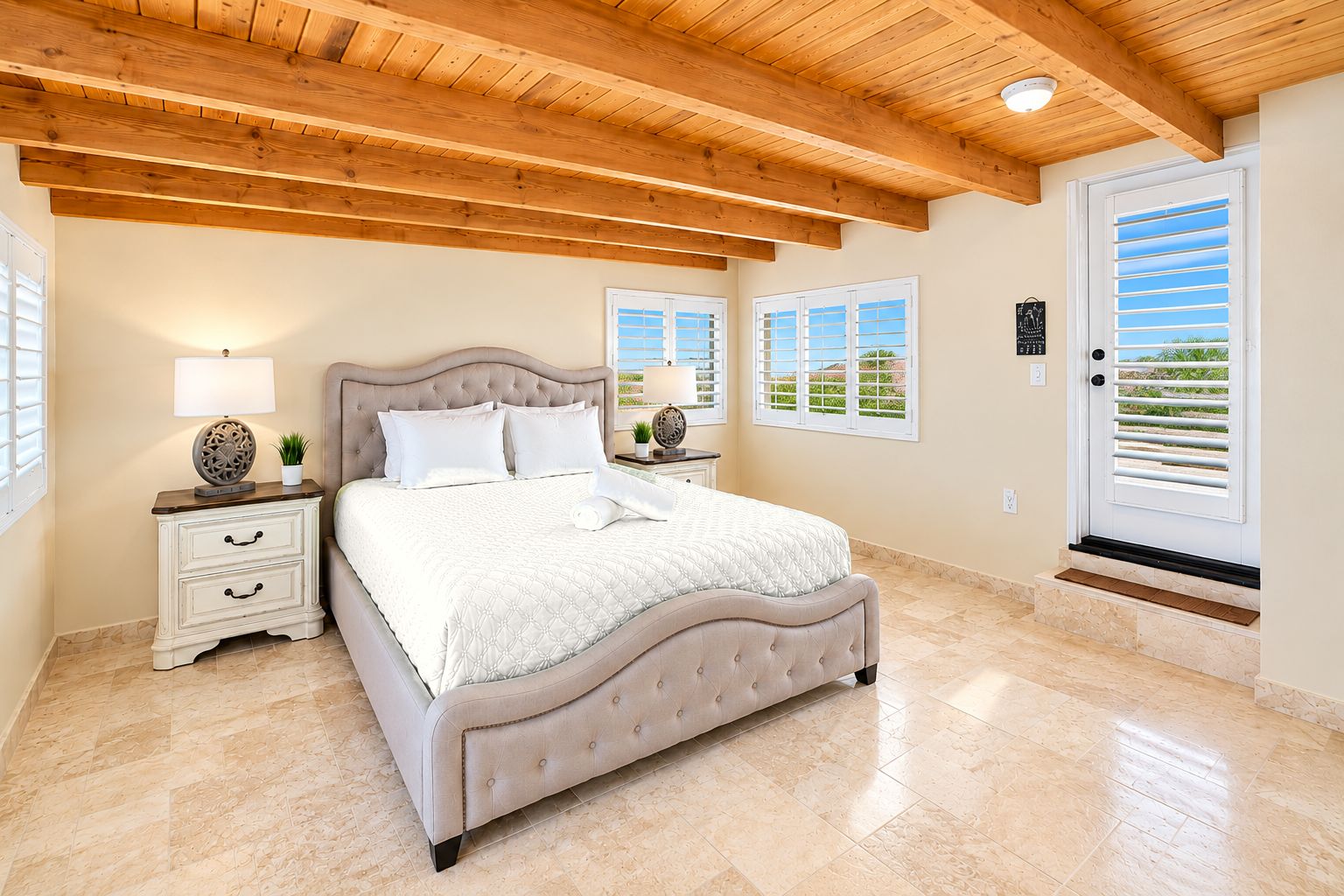 A bedroom with a large bed, wooden ceiling beams, a nightstand with a lamp, and a window with blinds.