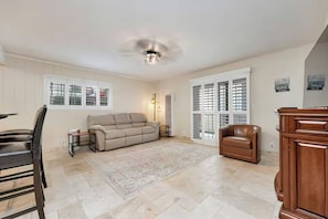 A spacious living room with a beige sofa, a brown leather chair, a dining table with chairs, a ceiling fan, and a large window with shutters. - 25% Off Oct 22-25, Steps To Sand, Roof-Top Deck! (Oxnard)