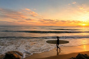 Une personne tenant une planche de surf sur une plage, avec des vagues et un coucher de soleil en arrière-plan.
