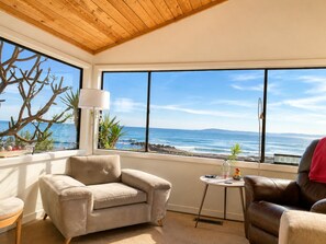 A room with a large window overlooking the ocean, a beige armchair, a small round table, and a brown leather chair. - 20% Off Sept! Water Front, Perfect For Families* (Ventura)