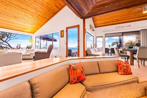 A modern interior with a wooden ceiling, a sofa with patterned cushions, and large windows offering a view of the sea.