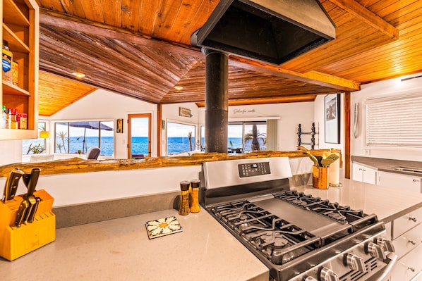 A kitchen with a gas stove, wooden ceiling, and a view of the sea through large windows.