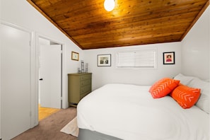 A bedroom with a wooden ceiling, a bed with white bedding, a dresser, and a window with blinds.