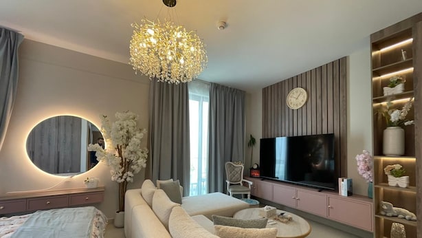 A modern living room with a large TV, a sofa, a round coffee table, and a decorative chandelier. - Stylish Studio Apartment with Kitchen, TV Lounge & Dining Space (Muḥāfaẓat al-Muḥarraq)